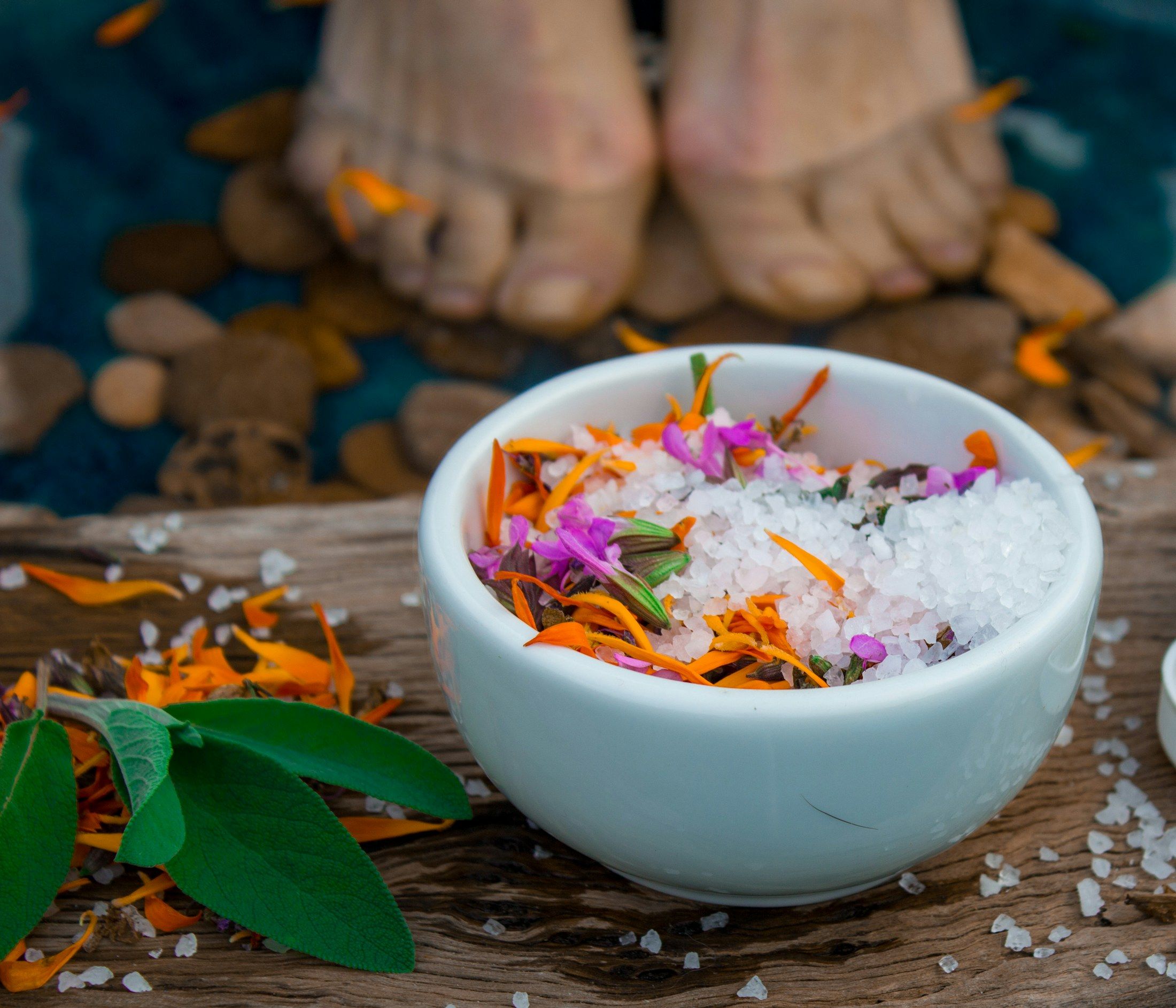 Schale mit Badesalz und Blüten, dahinter zwei Füße auf Kieseln